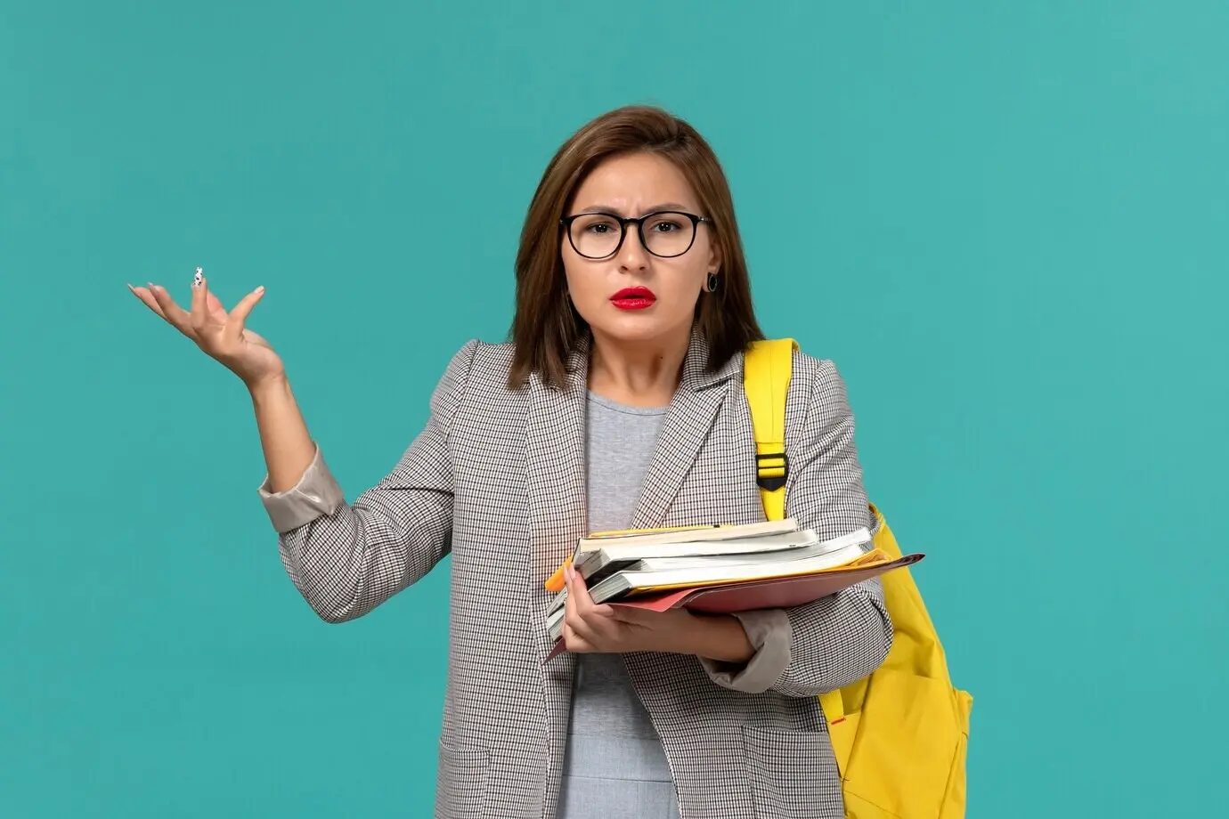 Vorderansicht einer Studentin in grauer Jacke mit gelbem Rucksack, die Bücher hält, vor einer hellblauen Wand.